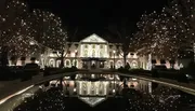 The image shows a large, stately house beautifully illuminated with white holiday lights, reflected in a calm body of water in front of it.