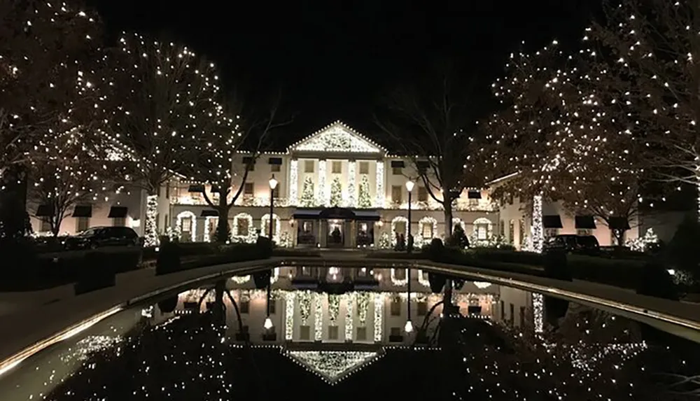 The image shows a large stately house beautifully illuminated with white holiday lights reflected in a calm body of water in front of it