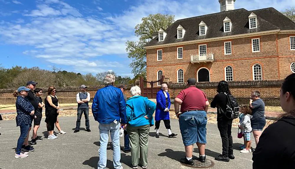 A group of people is gathered outdoors for a guided tour in front of a historic brick building under a partly cloudy sky