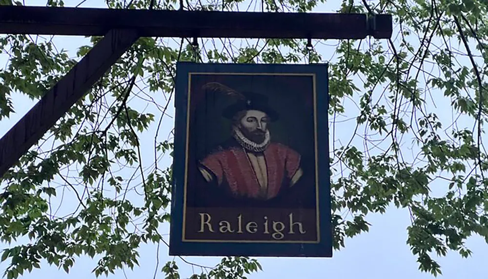The image shows a hanging sign featuring a painted portrait of a historical figure with the name Raleigh below set against a background of foliage and a clear sky