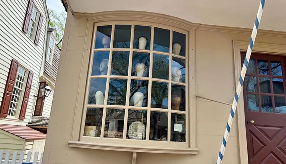 A bay window of a beige building displays multiple wigged mannequin heads overlooking a historical street with traditional wooden houses