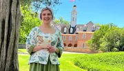 A smiling person in historical attire stands in front of a colonial-style building with an American flag.