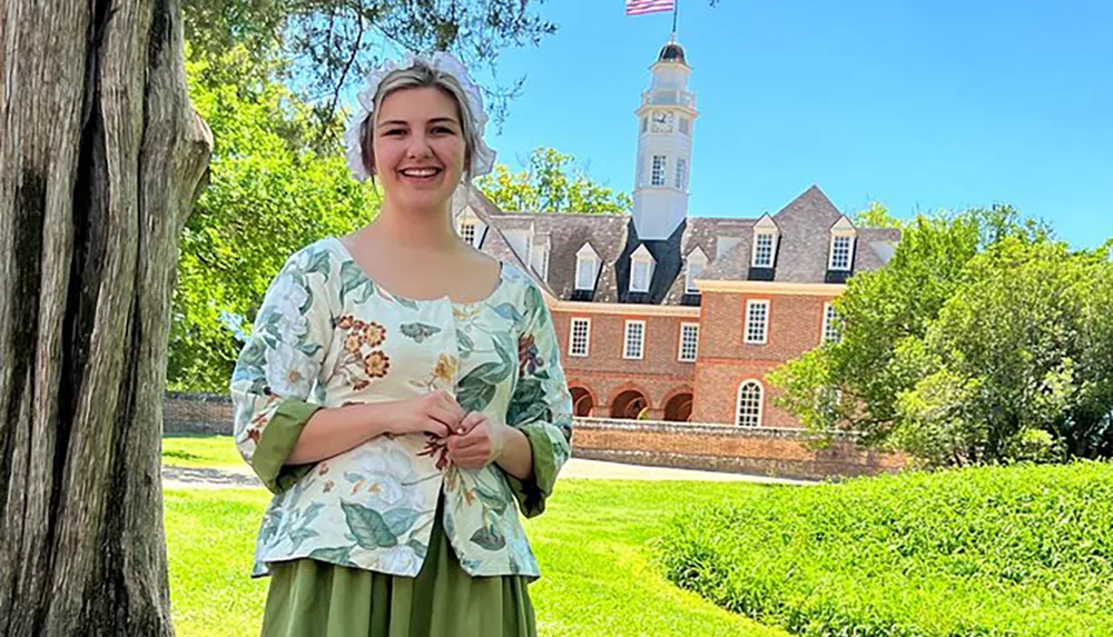 A smiling person in historical attire stands in front of a colonial-style building with an American flag