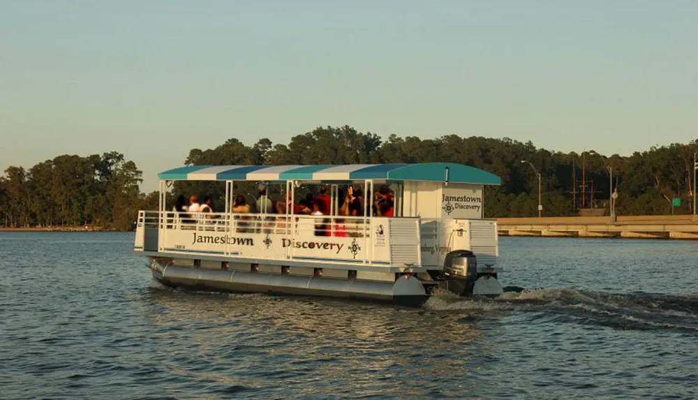 A ferry named Jamestown Discovery is cruising on the water with passengers on board during what appears to be late afternoon