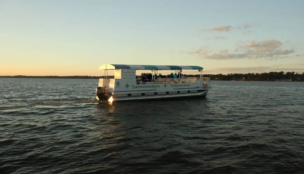 A pontoon boat named Jamestown Discovery is cruising on a calm body of water during sunset