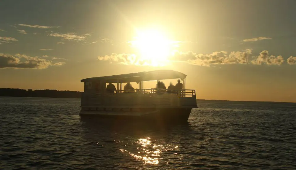 A pontoon boat carrying several passengers is silhouetted against a dazzling sunset over the water