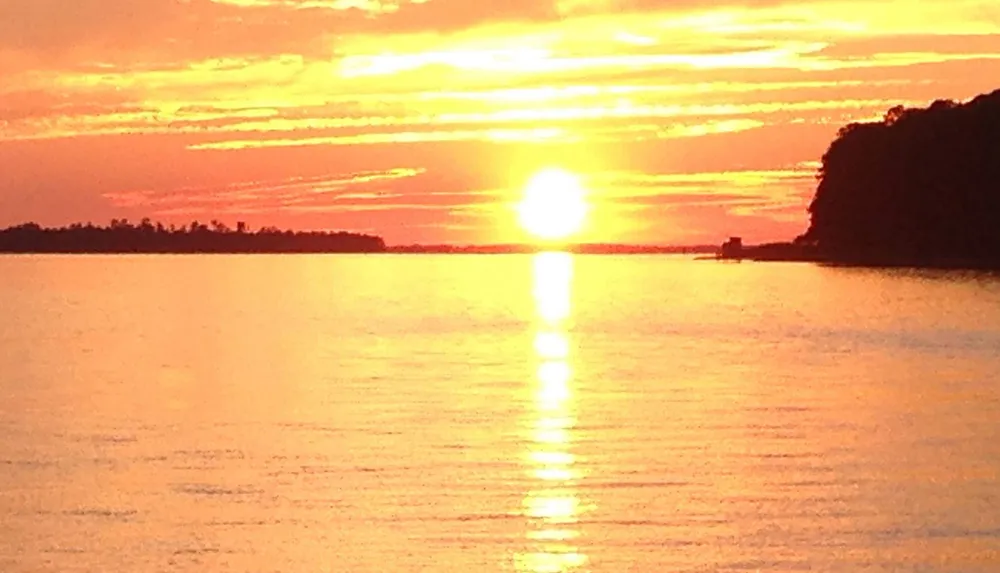 The image captures a serene sunset with the sun casting a golden glow across the sky and reflecting on the calm surface of the water flanked by silhouettes of land on either side