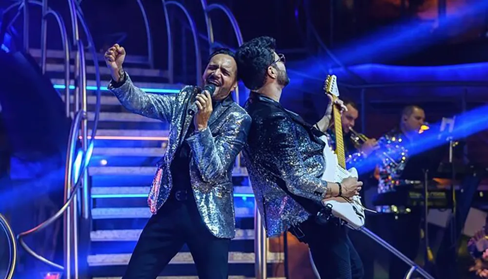 Two musicians in sparkling jackets are performing on stage with one singing passionately into a microphone and the other playing an electric guitar backed by band members under blue stage lighting