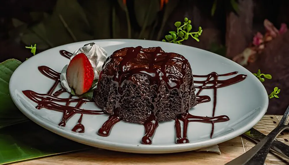 A delectable chocolate molten lava cake garnished with a sliced strawberry and dollop of cream is served on a white plate adorned with chocolate drizzle