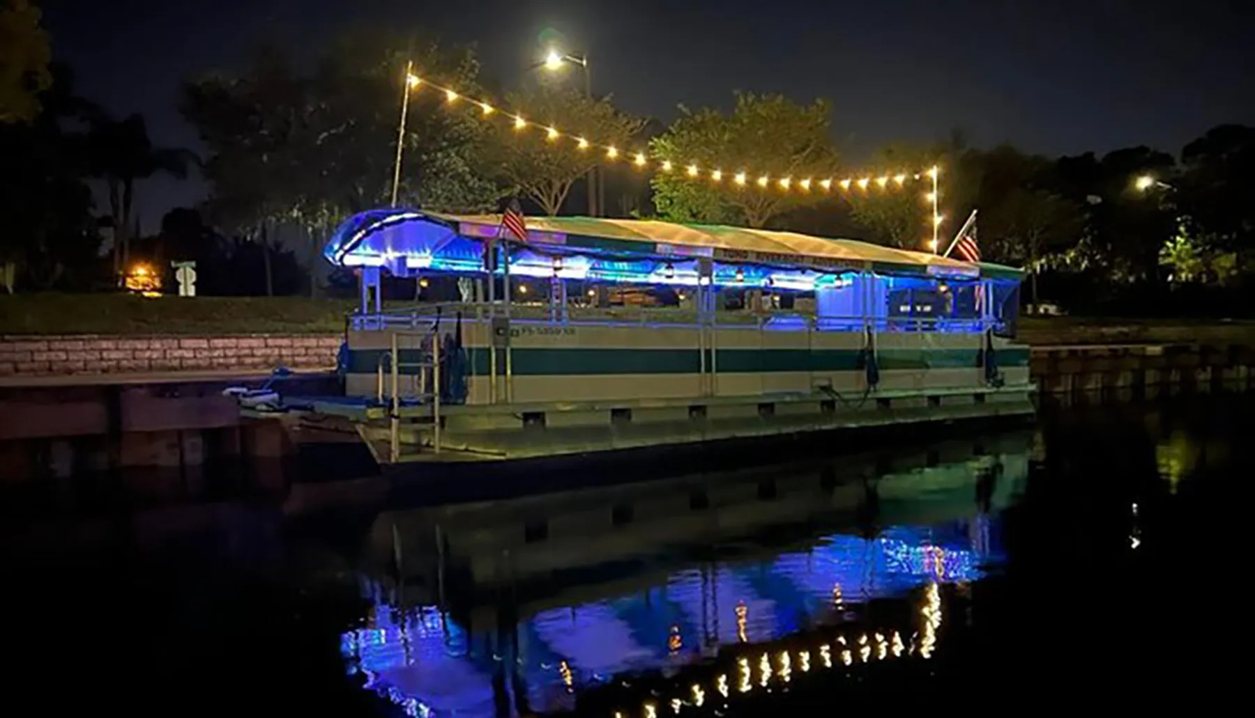 A boat decorated with lights is moored at a dock during the evening, reflecting in the water.