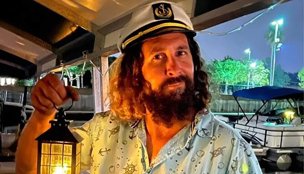 A man with a beard is holding a lantern and wearing a captains hat looking pleased with a marina in the background