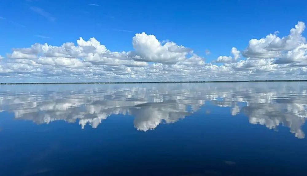 The image depicts a tranquil lake with a mirror-like surface creating a perfect reflection of the blue sky and fluffy white clouds above