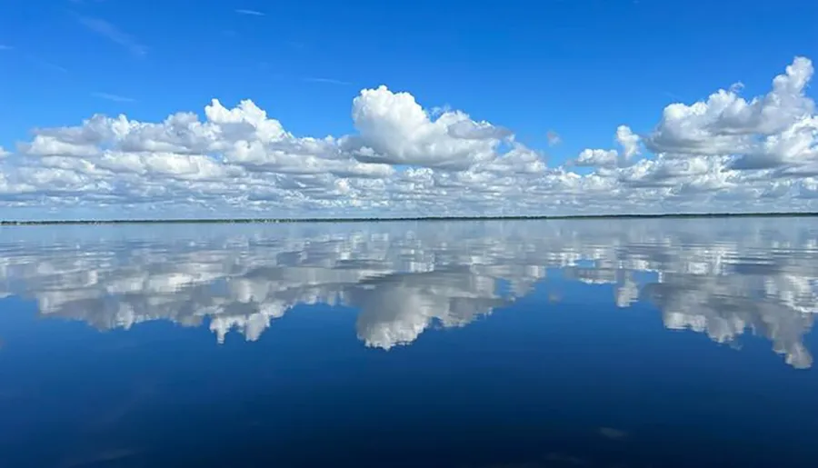 The image depicts a tranquil lake with a mirror-like surface creating a perfect reflection of the blue sky and fluffy white clouds above.