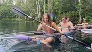 A group of individuals is kayaking in clear water, with one person in a transparent kayak revealing the underwater view.