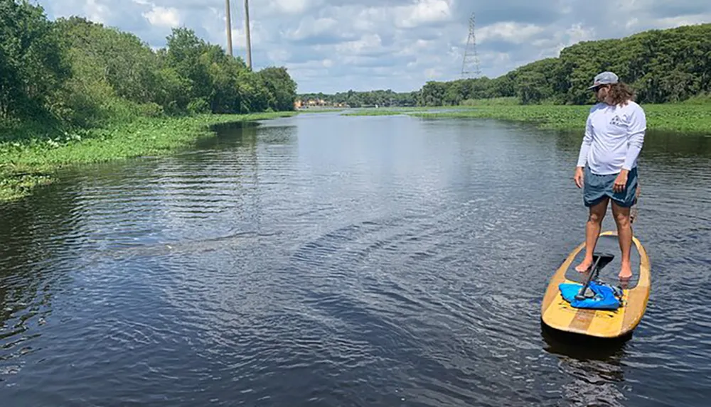 A person stands on a paddle board in a calm river surrounded by greenery under a cloudy sky