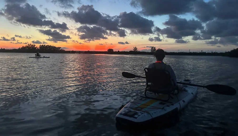 A person kayaks on calm waters against the backdrop of a stunning sunset with vivid orange hues painting the sky