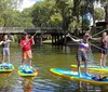 A group of people are standing on paddleboards in a waterway each holding a paddle aloft with a lush green backdrop and a bridge in the background