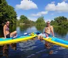 A group of people are standing on paddleboards in a waterway each holding a paddle aloft with a lush green backdrop and a bridge in the background