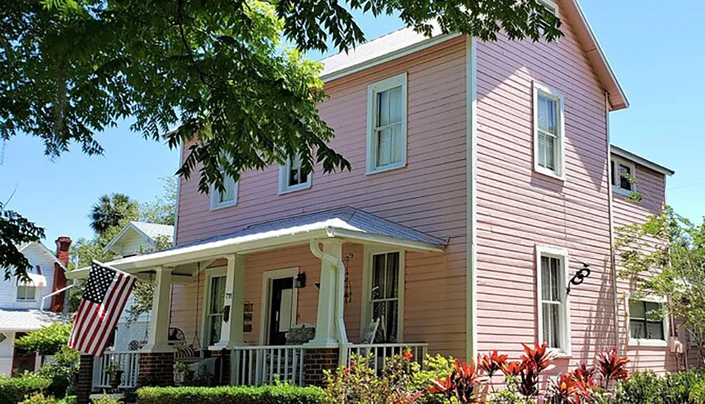 A quaint two-story pink house adorned with an American flag featuring a covered porch and surrounded by greenery under a clear blue sky