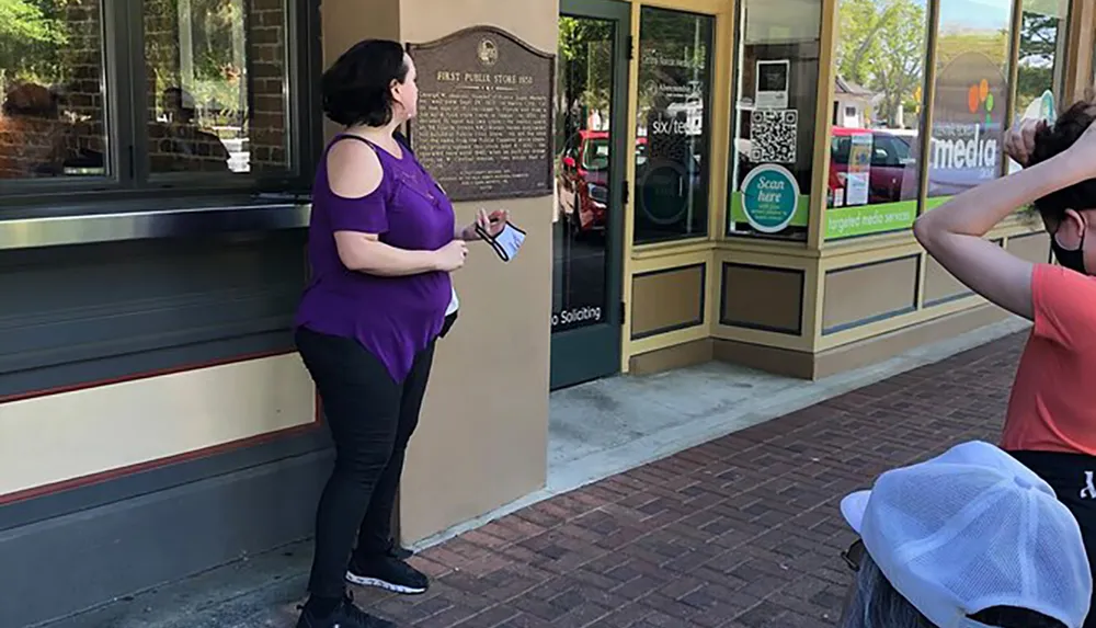 A woman in purple is standing on a sidewalk reading a plaque next to a building while another person in a red top appears to be adjusting their hair seated on the sidewalk