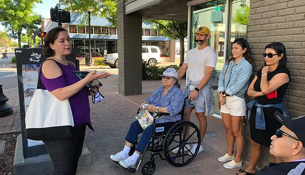 A group of people including one individual in a wheelchair is listening to a woman who appears to be explaining something outdoors