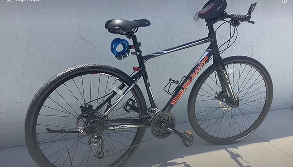 The image shows a black and orange mountain bike leaning against a gray wall with a blue bike lock wrapped around the frame and a black saddlebag attached to the seat