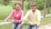 Two smiling people wearing cycling helmets are riding bicycles together in a park-like setting.