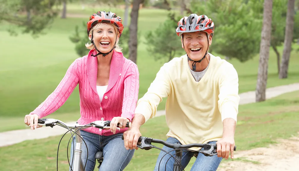 Two smiling people wearing cycling helmets are riding bicycles together in a park-like setting