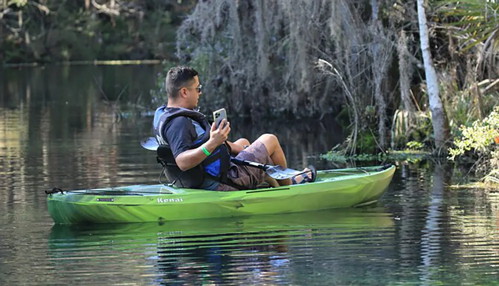 A person is sitting in a green kayak on calm water looking at a smartphone