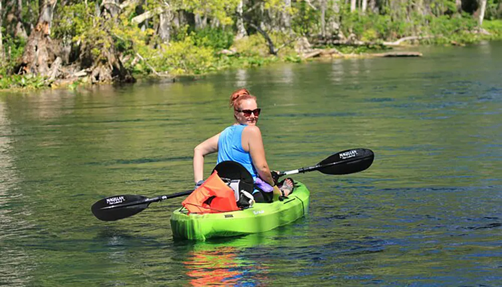 A person is kayaking on a clear and calm body of water surrounded by natural scenery