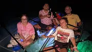 Four people are smiling and posing at night on a transparent canoe with underwater lighting that reveals the sea life below.