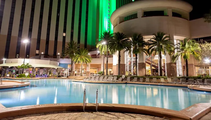 An illuminated swimming pool area with lounge chairs and palm trees is set against the backdrop of a sleek high-rise building at night