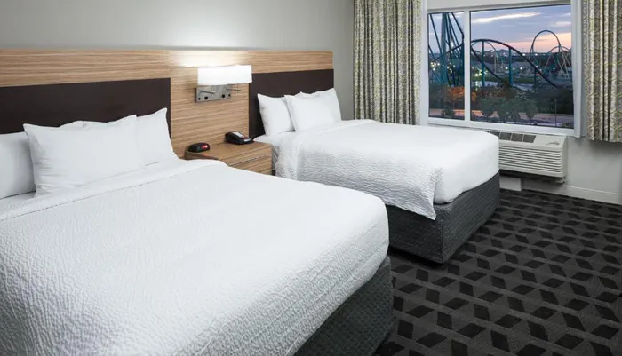 The image shows a modern hotel room with two neatly made beds and a large window that offers a view of a roller coaster and a ferris wheel outside