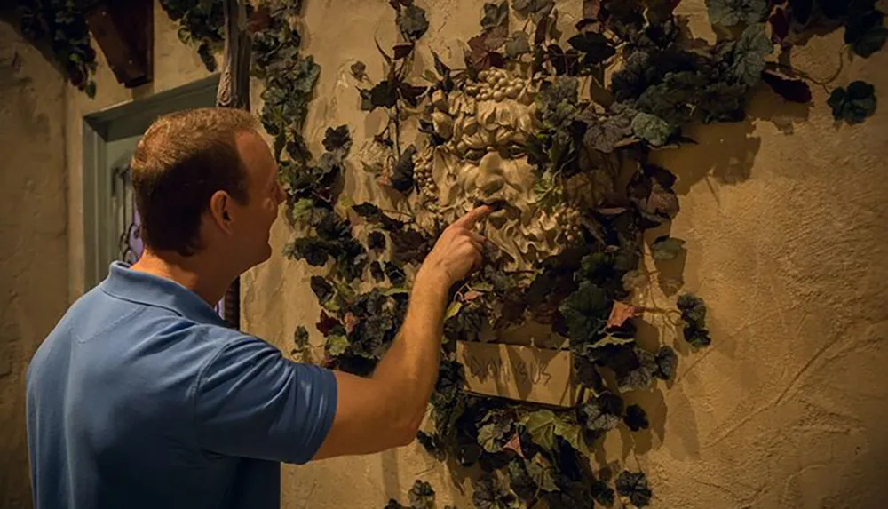 A man is touching a decorative piece featuring a face amidst foliage on a wall