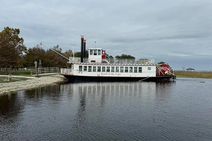 A paddlewheel boat named Pearl of the Lake is docked at a calm lakeside with an overcast sky above.