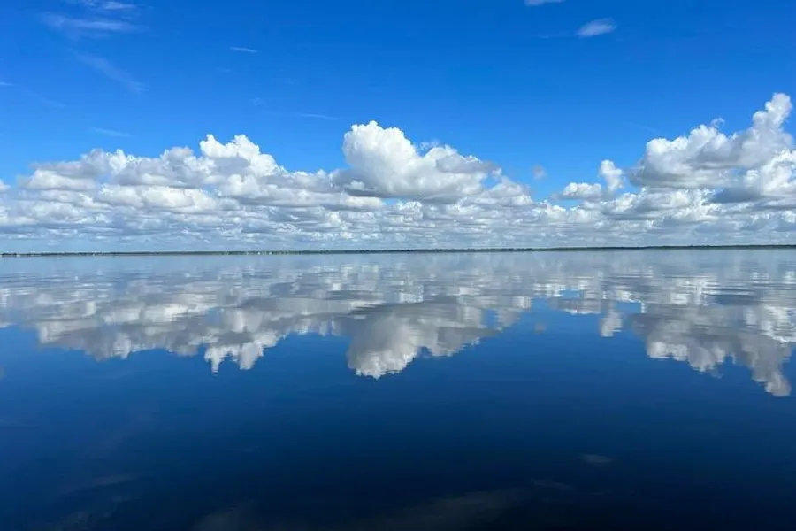 The image features a calm body of water with a clear sky above, where the white, fluffy clouds are perfectly reflected on the water's mirror-like surface, creating a serene and symmetrical natural scene.
