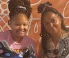 Two individuals are smiling for the camera while holding decorated sneakers standing in front of an orange wall with white line patterns