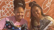 Two individuals are smiling for the camera while holding decorated sneakers, standing in front of an orange wall with white line patterns.