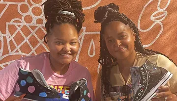 Two individuals are smiling for the camera while holding decorated sneakers, standing in front of an orange wall with white line patterns.