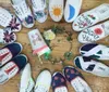 A variety of canvas shoes with different hand-painted designs are arranged in a circle around flowers and a notebook on a wooden floor