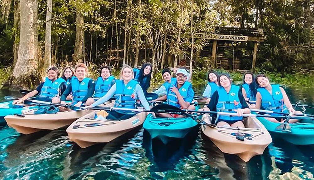 A group of people wearing life jackets are smiling for a photo while seated in a flotilla of kayaks on a clear water body near a dock named ESP SPRINGS LANDING
