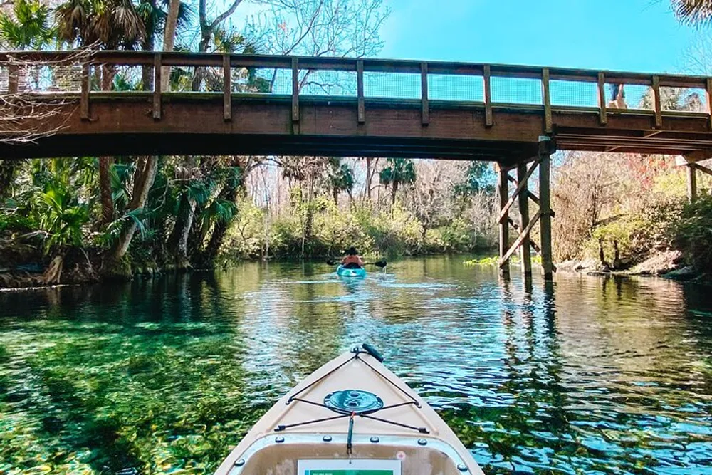 A kayak floats on clear water under a wooden bridge with lush greenery on a sunny day