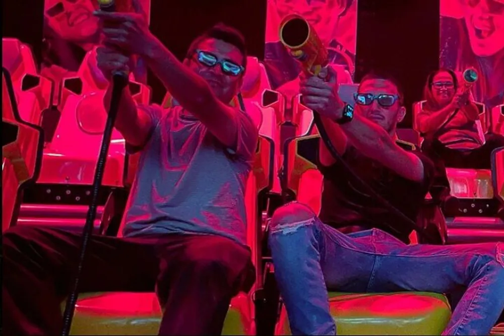 Two individuals are wearing 3D glasses and holding toy guns in a vibrant red-lit arcade shooting game area appearing to be engaged in an immersive gaming experience