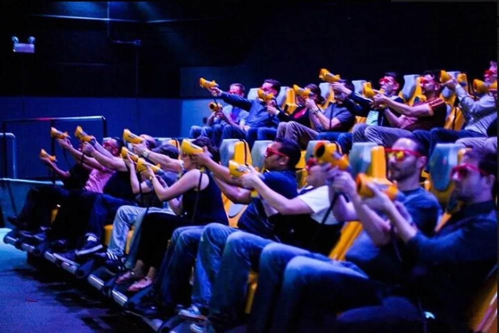 A group of people is seated in a dark room each aiming a yellow toy gun as part of an interactive theater or gaming experience