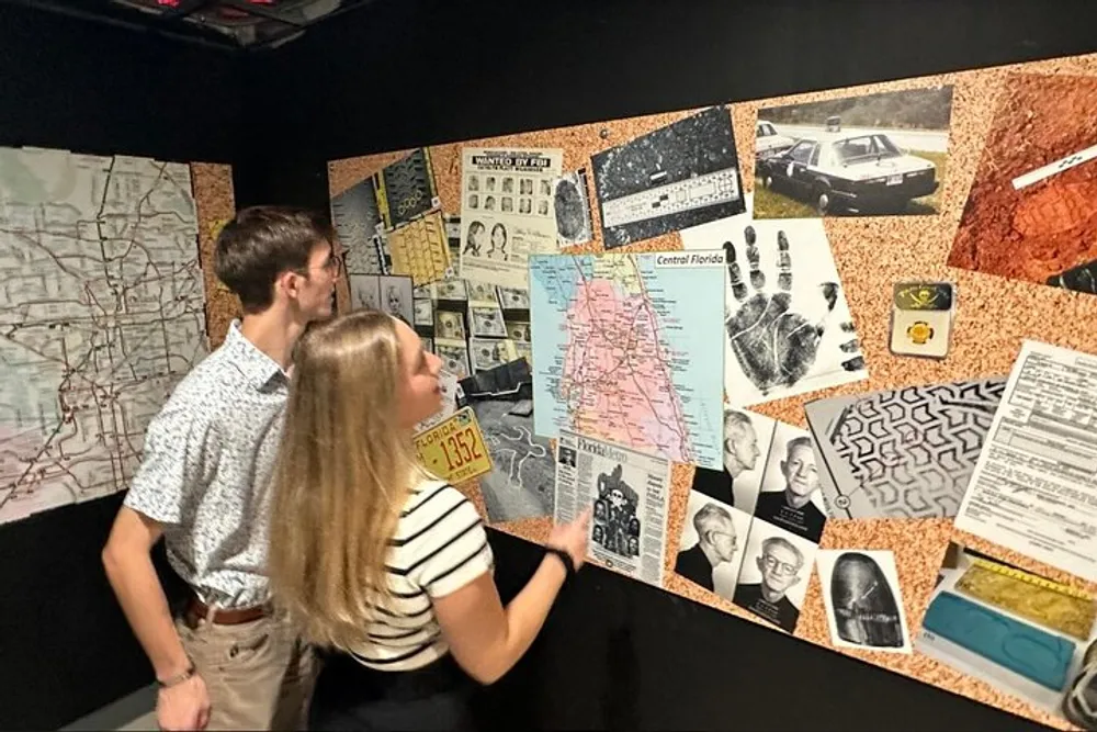 Two people are examining a bulletin board filled with various papers photos and maps possibly reviewing details of a case or project