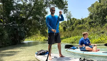Two people are enjoying paddleboarding on a calm river surrounded by lush greenery on a sunny day.