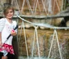 A young girl with a sunny smile holds onto the ropes of a rope bridge in front of a softly blurred waterfall background