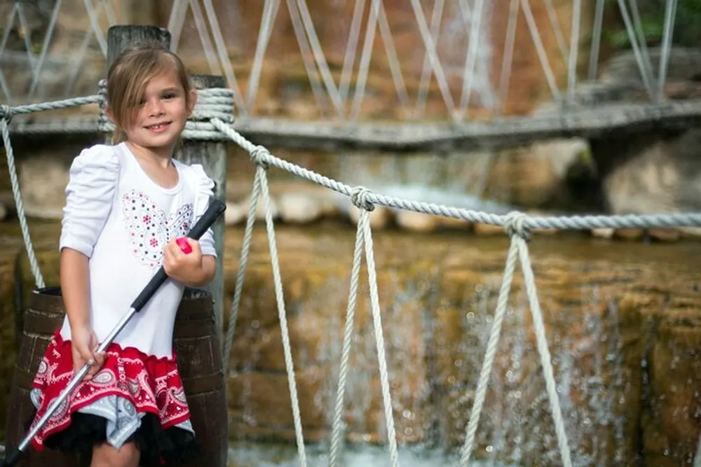 A young girl with a sunny smile holds onto the ropes of a rope bridge in front of a softly blurred waterfall background