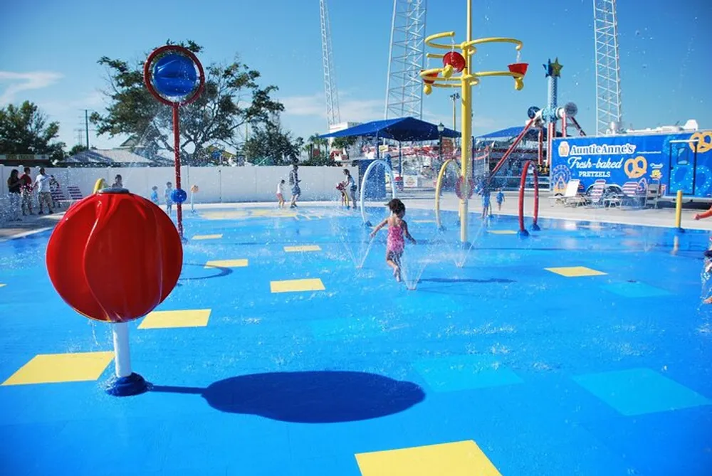 The image shows children playing in a vibrant water park with colorful splash pads and water features on a sunny day