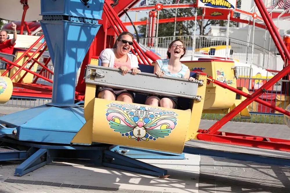 Two people are joyfully screaming while riding a colorful amusement park ride on a sunny day
